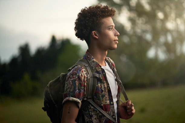 Outdoor portrait of young African-American backpacker Waist-up profile view of contemplative man with curly brown hair wearing casual clothing, backpack, and looking away from camera at dusk. man standing profile stock pictures, royalty-free photos & images