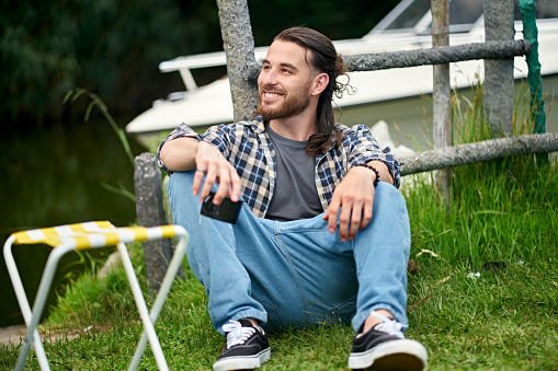 Full length front view of smiling bearded man in casual clothing sitting on ground and looking away from camera while enjoying weekend getaway.
