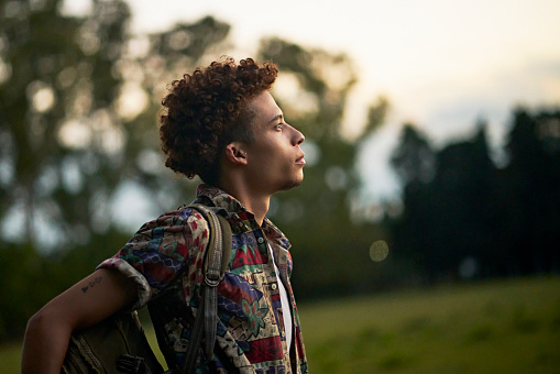Waist-up profile view of man with curly brown hair wearing casual clothing and backpack looking away from camera with contented expression of hope.