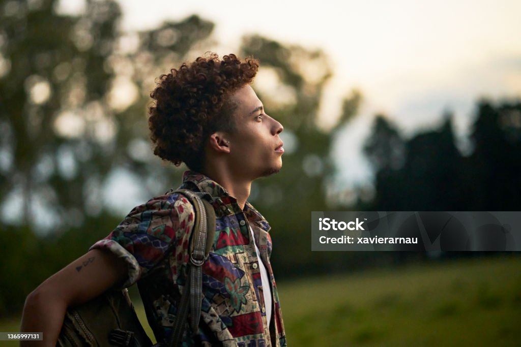 Portrait of mid 20s African-American man outdoors at dusk Waist-up profile view of man with curly brown hair wearing casual clothing and backpack looking away from camera with contented expression of hope. Contemplation Stock Photo