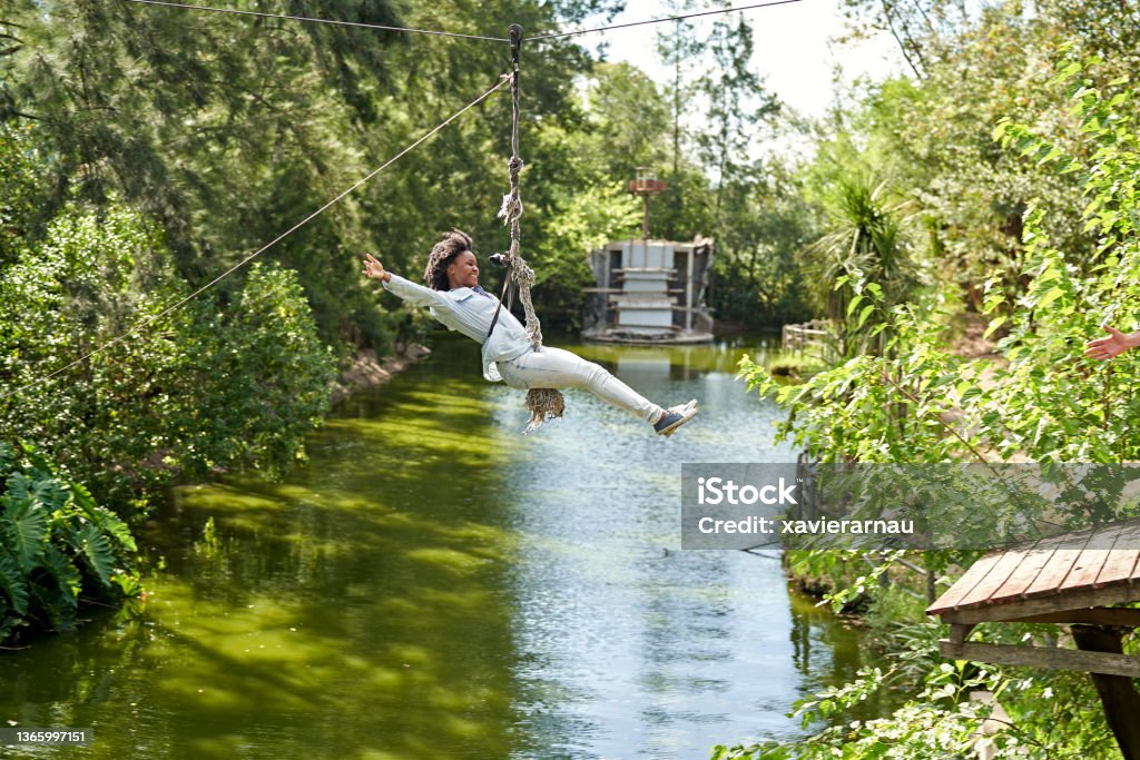 Smiling young Black woman riding zip line across pond Full length side view of woman in casual clothing hanging in mid-air from pulley with arms outstretched and legs extended. Zip Line Stock Photo