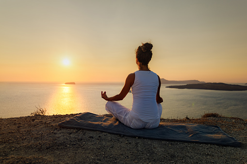 Rear view of a relaxed woman meditating in Lotus position on a hill above the sea at sunset. Copy space.