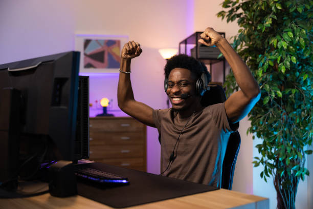 istock Winning a round in a computer video game. A dark-skinned man with a headset shouts to the team for the joy of victory, a gamer clenches fists in triumph, the room dark with colored led lights 1366001048