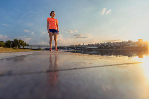Young woman in running shorts standing on the platform by the river in the city at dusk, lifestyle in motion