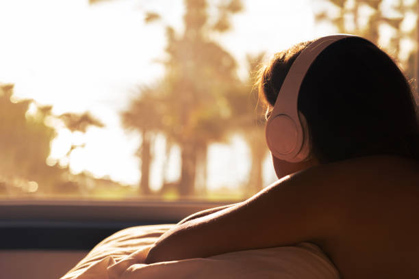 silhouette de femme avec des écouteurs allongée sur un oreiller sur le lit dans la chambre en face de la fenêtre panoramique avec vue sur la plage de la mer de palmier au coucher du soleil. femme relaxante écoutant de la musique. - tree house audio photos et images de collection