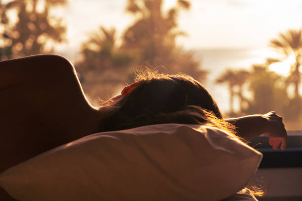 Silhouette of woman lying on the bed and sleeping on pillow in bedroom opposite panoramic window with view on palm tree sea beach at sunset. Back view. stock photo