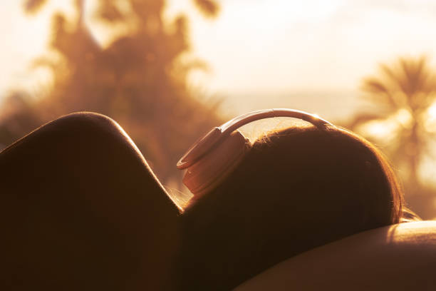 sylwetka kobiety ze słuchawkami leżącej na poduszce na łóżku w sypialni naprzeciwko panoramicznego okna z widokiem na palmową plażę morską o zachodzie słońca. kobieta relaksująca słuchająca muzyki. - tourist resort audio zdjęcia i obrazy z banku zdjęć