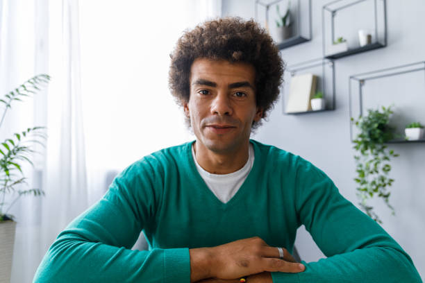 Confident m ultiracial man smiling at camera as if h Front view of young multiracial man smiling at camera as if he is on a video conference while working from home. man video call pov stock pictures, royalty-free photos & images