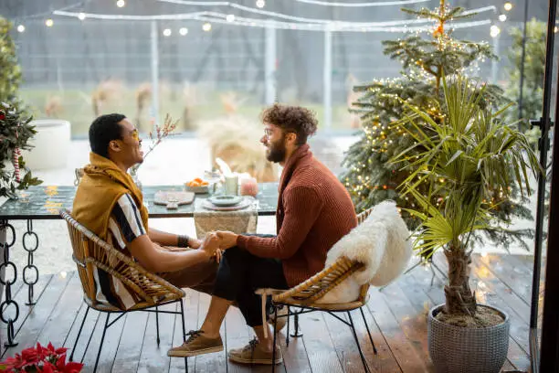 Two multiracial male friends having close conversation, sitting by a dinner table on a beautiful house terrace outdoors. Caucasian and hispanic man together at backyard