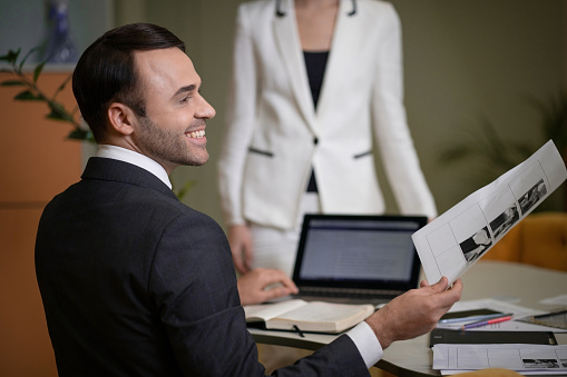 Businessman in a Classic Suit and White Shirt Sits at his Desk in Profile to the Camera. Head of the Company Delegates Tasks in his Office. White Collar Worker .In the Background is a Business Lady in a White Formal Suit. Burred Background. Close-up. High quality photo