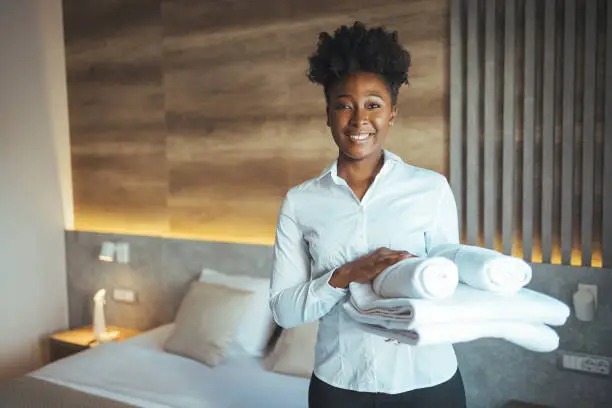 Maid working at a hotel holding towels and looking at the camera smiling - housekeeping concepts. Maid with fresh clean towels during housekeeping in a hotel room