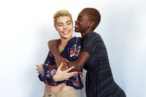 Photo of joyful women kissing and embracing togetherness, being of different of races, dressed in casual clothes.