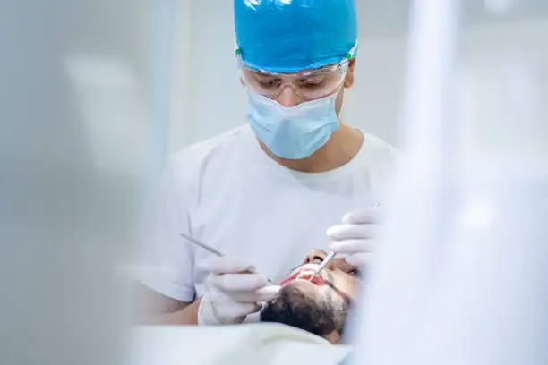 Handheld shot of a dentist with a protective face mask, medical gloves, and surgical cap, using a mouth mirror and dental probe, to exam teeth and gums, of a male African-American patient.