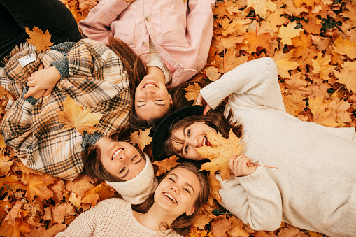 Top view of young smiling fair-haired and dark-haired women friends, two of them having hats on their heads, dressed in white, grey, pink and beige jackets, lying in orange autumn leaves in park