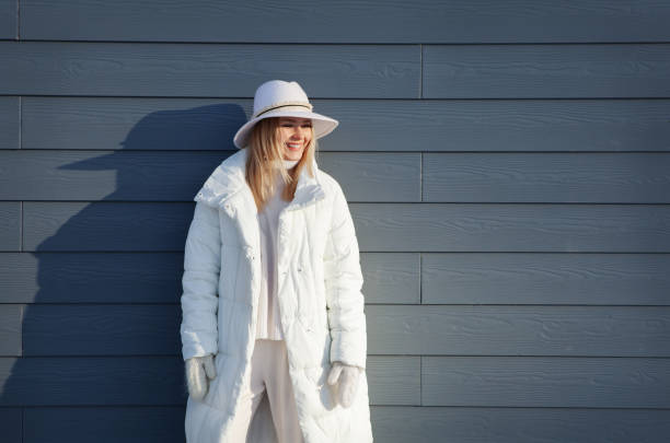 太陽からの影を持つ灰色の背景に笑うブロンドの女性の本物の肖像画、冬の暖かい白い服で目をそらし、幸せな顔の表情 - coat ストックフォトと画像