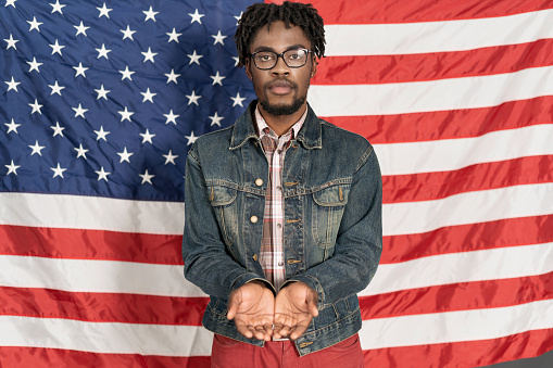 Poor African American Asks for Money from the State. Guy Stands with Folded Hands Begging in the US Flag Background .Life on Unemployment Benefits. Poor Immigrant. Close-up .Copy Space. High quality photo