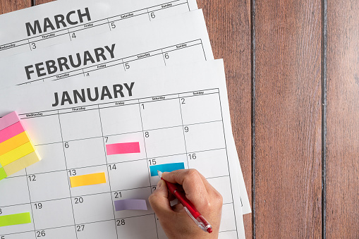 View from above of a person's hand making notes on a calendar with the first months of the year using colored post it notes to highlight the activities. Concept plans and objectives, fulfillment of goals. High quality photo