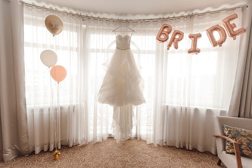 Gorgeous white wedding dress hanging on hanger against window in sunlit room decorated with white, pink and golden balloons. Bride's morning gathering and preparation