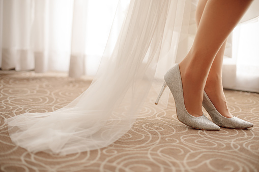 Crop photo of unrecognisable bride's veil and legs demonstrating stiletto heeled shoes, standing indoors on beige flooring near window while preparing for wedding day. Bride's morning gathering