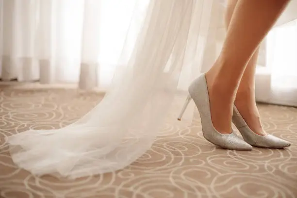 Photo of Crop photo of unrecognisable bride's legs demonstrating stiletto shoes standing indoors on beige flooring near window