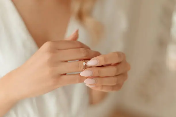 Crop photo of unrecognisable young bride's hands with light manicure, wearing white morning gown and putting on wedding ring on finger while preparing for her wedding day. Bride's morning gathering