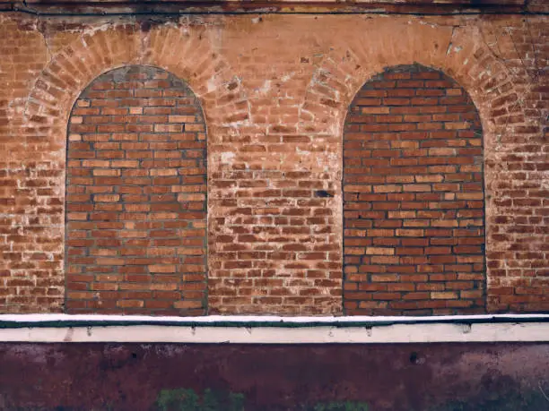 The brick wall of the old building, the arched window openings are bricked up, the paint on the wall has burned out, and in places it has peeled off, the old texture
