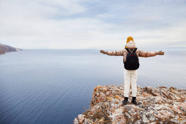 una giovane ragazza viaggiatrice in abiti caldi con uno zaino è in piedi sulla scogliera, ammirando la natura. gode della vista sul mare o sul lago nel tardo autunno. - lake baikal lake landscape winter foto e immagini stock