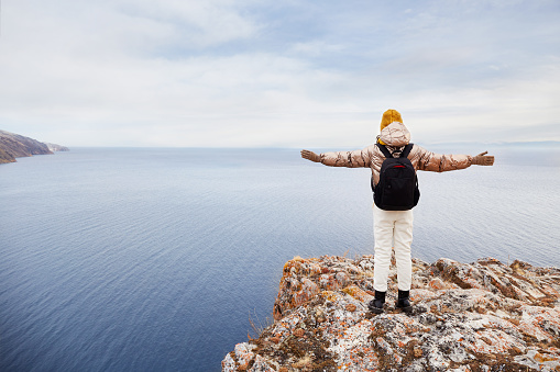 A young girl traveler in warm clothes with a backpack is standing at the cliff, admiring nature. Enjoys the view of the sea or lake in late autumn. Traveling alone, healthy lifestyle, freedom.