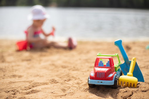 A toy truck is standing with a shovel and a rake on the beach, in the background a beautiful girl in a white panama hat is playing in the sand