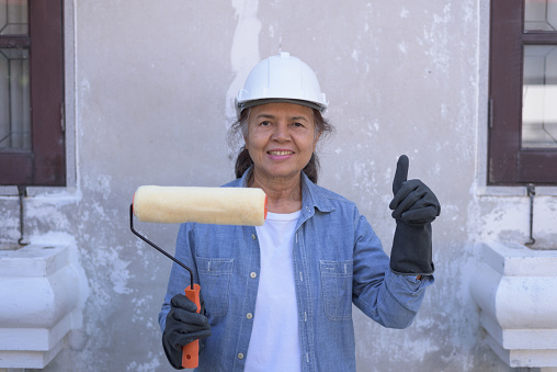 Front view, portrait of a happy Asian female worker wearing white safety hard hat and gloves, holding paint roller, smiling, putting thumb up. Retired senior lifestyle and DIY Home renovation concept.