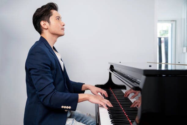 파란 양복과 청바지를 입은 자신감 넘치는 남자가 의자에 앉아 흰색 배경을 가진 그랜드 피아노를 연주합니다. 기대. 뮤지션은 멜로디를 재생합니다. 초상화 및 라이프스타일 컨셉 - pianist 뉴스 사진 이미지