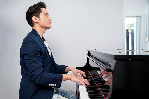 A confident man wearing blue suit and jean sitting on the chair and playing grand piano with white background. Looking forward. Musician play melody. Portrait and lifestyle concept