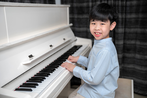Asia boys wearing shirt look at camera and smile playing white piano. Enjoying time practicing music at school.  Musician kid feeling happy and relax during class. Education and learning concept