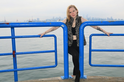 A woman posing with a blue railing next to Vancouver BC Canada harbor. She is wearing a black top, pants, shoes and a leopard print sleeveless vest.