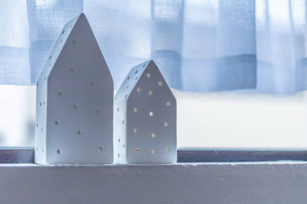 Two white decorative miniature houses on a windowsill. White courtain in background.