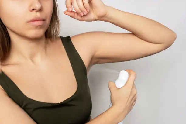 Cropped shot of a young caucasian blonde woman applying a spray antiperspirant to the skin of the armpits isolated on a gray background. Deodorant for masking and blocking the smell of sweat