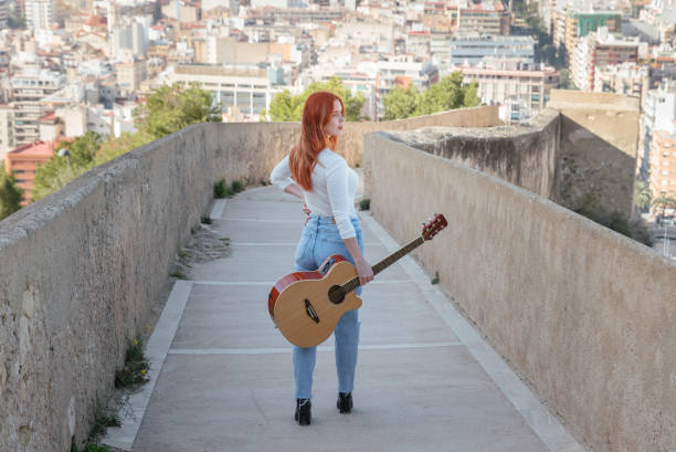 istock Beautiful young redhead walks with her guitar outdoors along a wall that has panoramic views of the city 1366106271