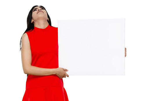 Front view of aged 20-29 years old who is beautiful with black hair latin american and hispanic ethnicity young women standing in front of white background wearing dress who is feeling sad who is showing with hand and holding banner sign with copy space