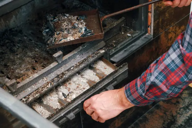 Photo of Ash removal from the fireplace. A man's hand in a plaid shirt holds a shovel with ash from the fireplace. A man cleans the fireplace.