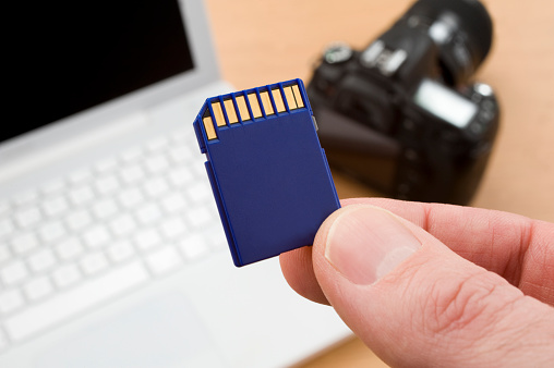 Close up of SD card with computer and camera in background.