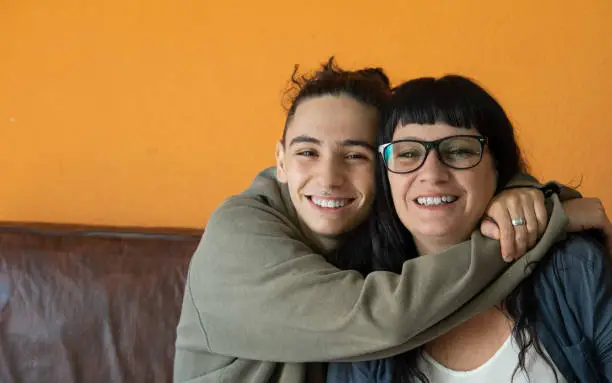 Photo of handsome teenage boy hugging his mother affectionately and smiling at the camera while sitting on the sofa. Mother and son. family concept.