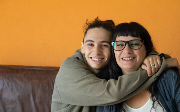 handsome teenage boy hugging his mother affectionately and smiling at the camera while sitting on the sofa. mother and son. family concept. - mid teens imagens e fotografias de stock