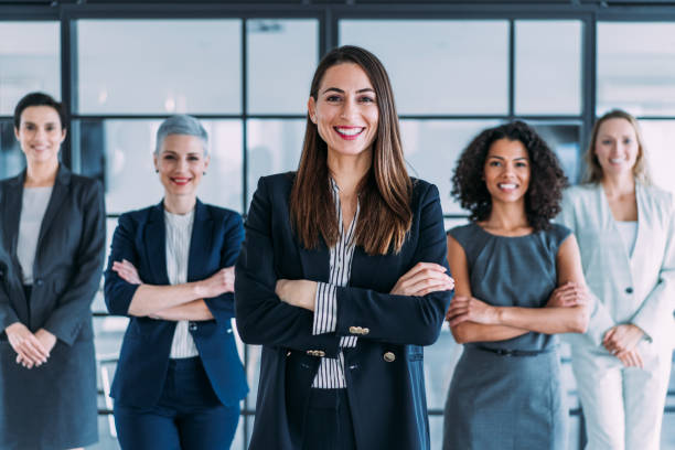 Confident businesswoman and her female team. Portrait of beautiful smiling businesswoman with her colleagues. Multiracial  group of businesswomen standing in the office. Successful team leader and her female team in background. Shot of five confident young businesswomen standing in modern office and looking at camera. Group of businesswomen standing together in office. formal businesswear stock pictures, royalty-free photos & images