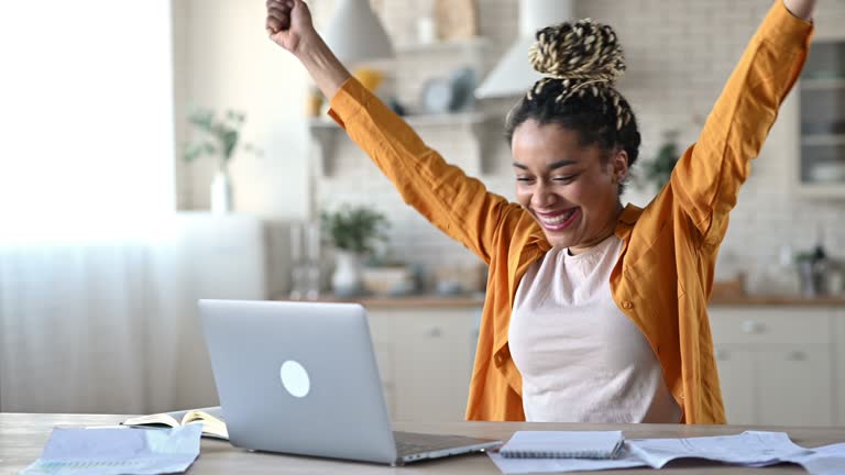 Overjoyed excited african american girl with dreadlocks, freelancer, manager working remotely at home using laptop, looks at screen with surprise, smiling face, gesturing with hands, got a dream job