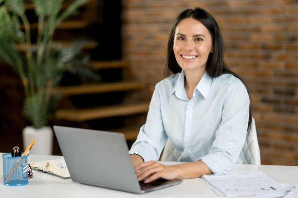 Portrait Caucasian female stock trader in office. Successful smart business woman, top manager or broker, brunette, sits in office, texting with colleagues and clients, taking notes, looking at camera Portrait Caucasian female stock trader in office. Successful smart business woman, top manager or broker, brunette, sits in office, texting with colleagues and clients, taking notes, looking at camera stock trader stock pictures, royalty-free photos & images