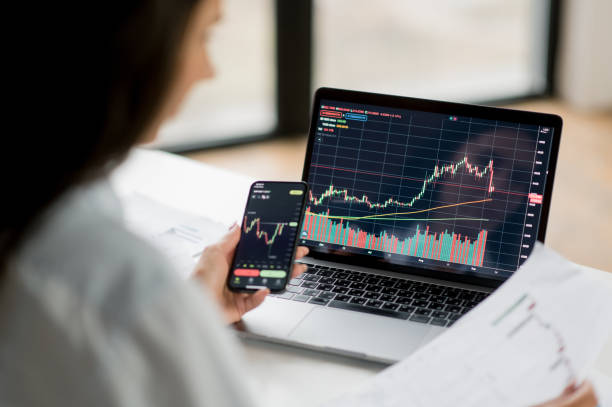 Successful smart woman investor and crypto trader, using laptop and smartphone, analyzes charts of trading in stock market and digital cryptocurrency exchange, conducts analysis, trading crypto coins stock photo