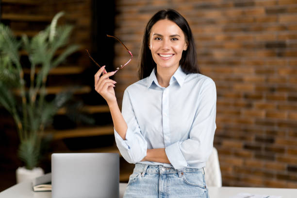 retrato feliz inteligente exitoso caucásico moreno mujer de negocios, ceo, alto gerente, agente de bienes raíces, se para cerca de la mesa en la oficina moderna, con ropa elegante, mira a la cámara, sonríe amigablemente - technology personal accessory hightech business fotografías e imágenes de stock