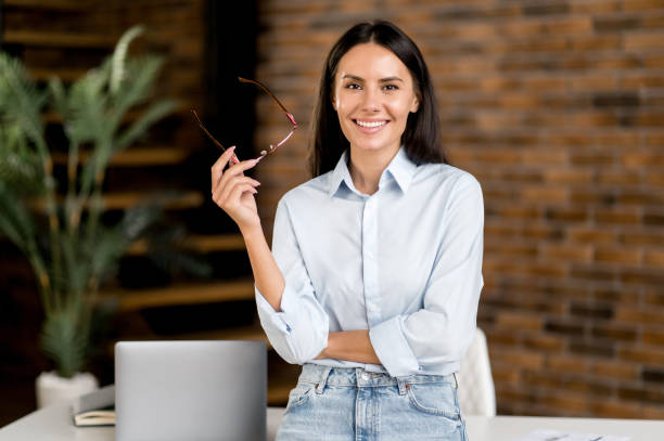 Portrait happy smart successful caucasian brunette business woman, ceo, top manager, real estate agent, stands near table in modern office, in stylish clothes, looks at camera, smiles friendly stock photo