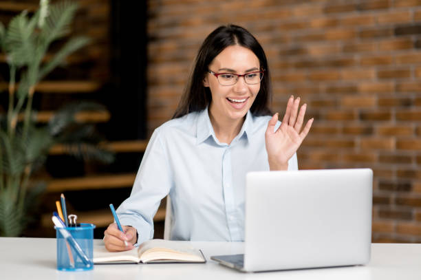 Friendly cute confident smart caucasian businesswoman, freelancer or manager wearing glasses, sitting at the table in the office, video communication with colleagues or client, waving hand, smiling Friendly cute confident smart caucasian businesswoman, freelancer or manager wearing glasses, sitting at the table in the office, video communication with colleagues or client, waving hand, smiling online tutors stock pictures, royalty-free photos & images
