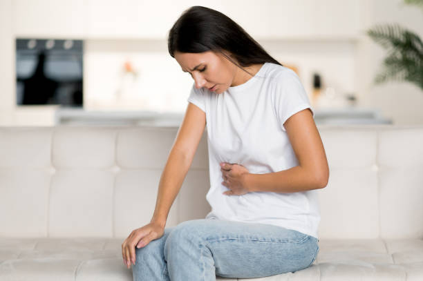 Frustrated young caucasian woman, sitting on the sofa in the living room, experiencing abdominal pain, health problems, cramps, ulcer, problems with the digestive tract, need a doctor's consultation stock photo